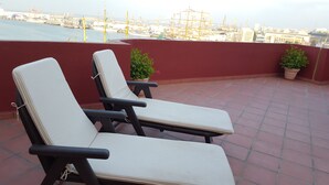 Deux fauteuils de jardin avec coussins, placés sur une terrasse carrelée donnant sur une marina où l’on peut voir des bateaux et des grues.