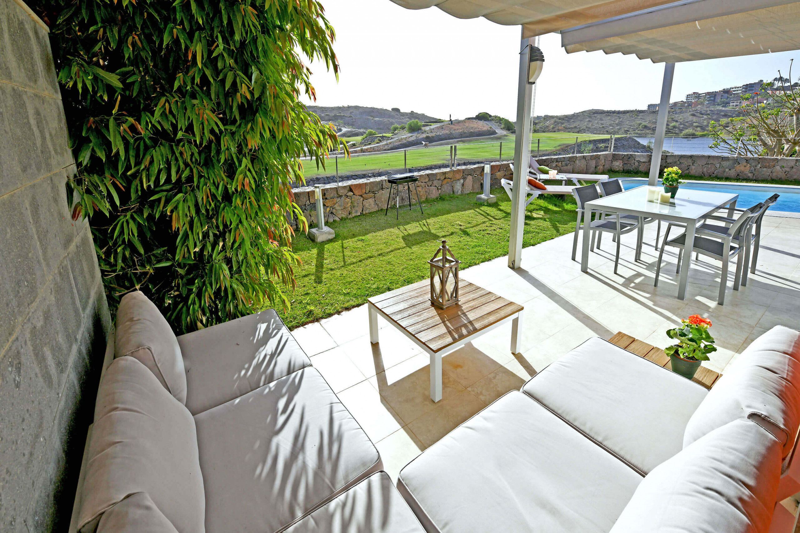 A patio with white outdoor furniture, a table, and a view of a swimming pool and hills.