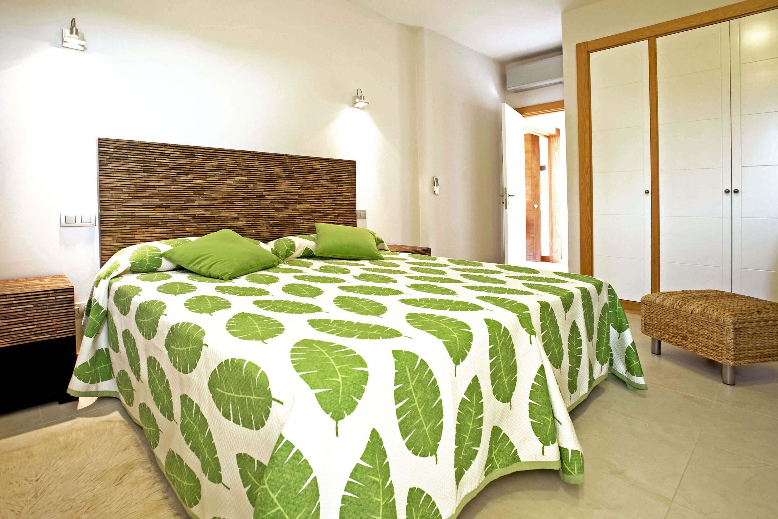 A bedroom with a large bed, green leaf-patterned bedspread, a wooden headboard, bedside tables, and a wicker bench.