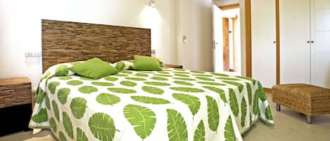 A bedroom with a large bed, green leaf-patterned bedspread, a wooden headboard, bedside tables, and a wicker bench.