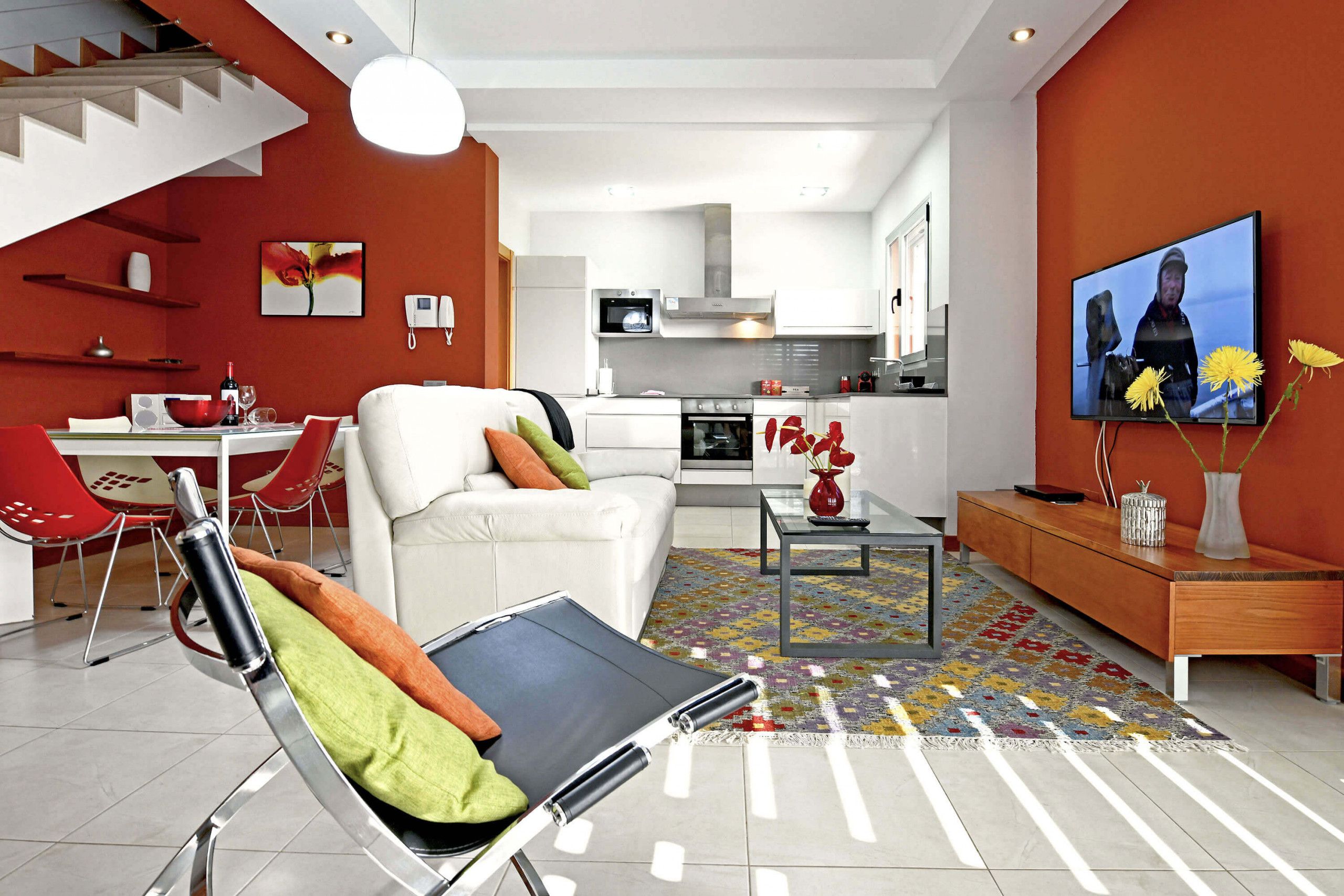 A modern living room with a sofa, a coffee table, and a TV. There is a kitchen in the background with white cabinets and a microwave.