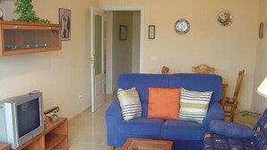 Un salon avec un canapé bleu, un coussin orange, une table basse en bois et un téléviseur posé sur un meuble en bois.