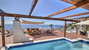 Zona de piscina con vista al mar, pérgola de madera y mobiliario exterior.