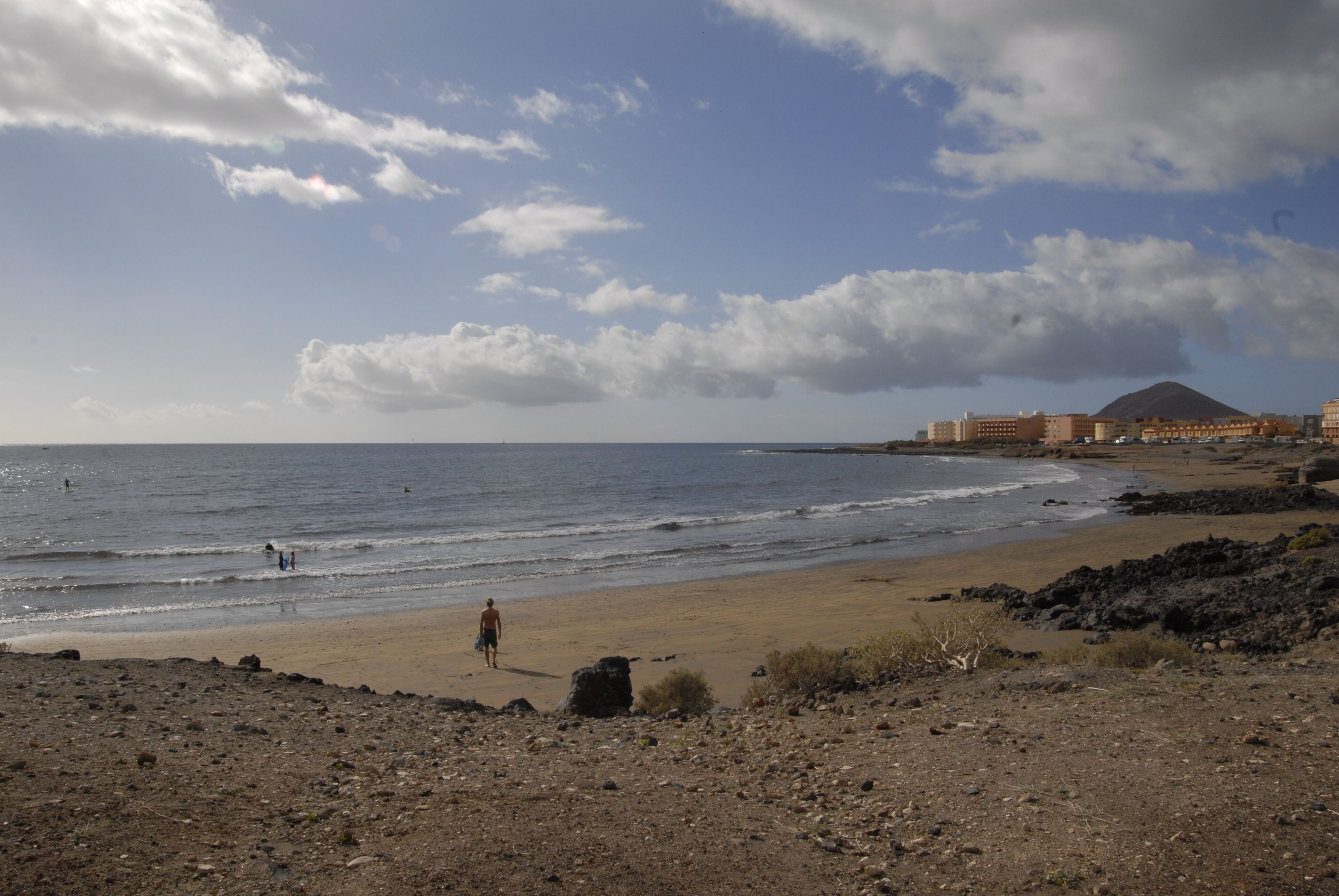 A beach with rocky terrain, a person walking, and buildings in the background.