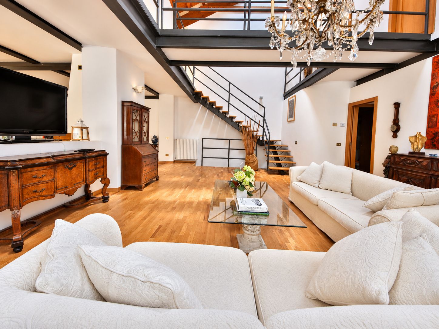 Ein modernes Wohnzimmer mit einem Glastisch, weißen Sofas, einem Holzschrank, einem Fernseher und einer Treppe mit schwarzer Geländer.