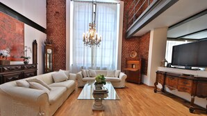 A spacious living room with a large window, a chandelier, a television, and a fireplace. - Loft by KlabHouse w/3BDRM+3BTH+Jacuzzi and Parking near Fond. Prada  (Milano)