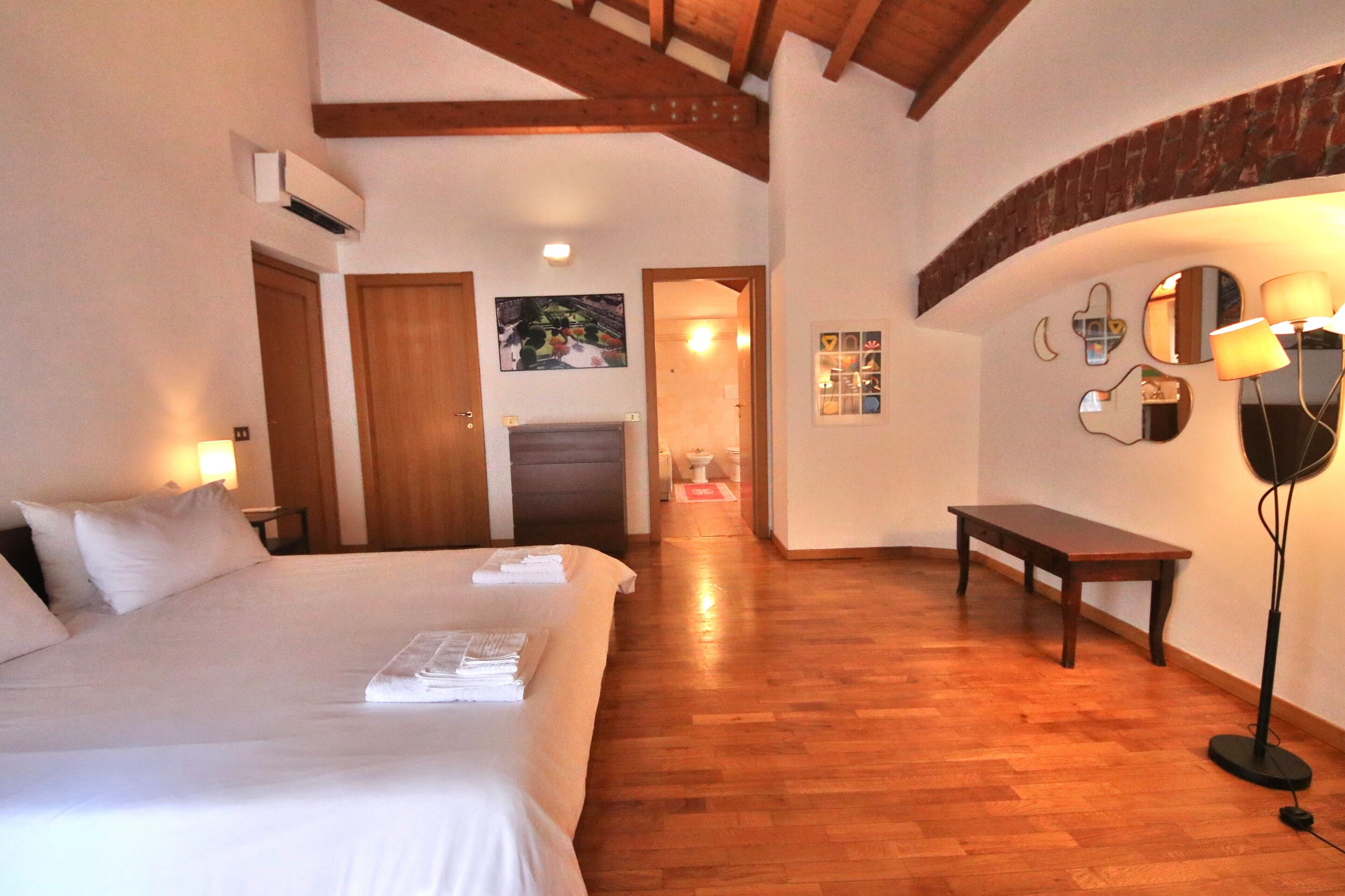 Quarto de hotel com uma cama grande, piso de madeira, um banco, espelho fixado na parede e lareira.