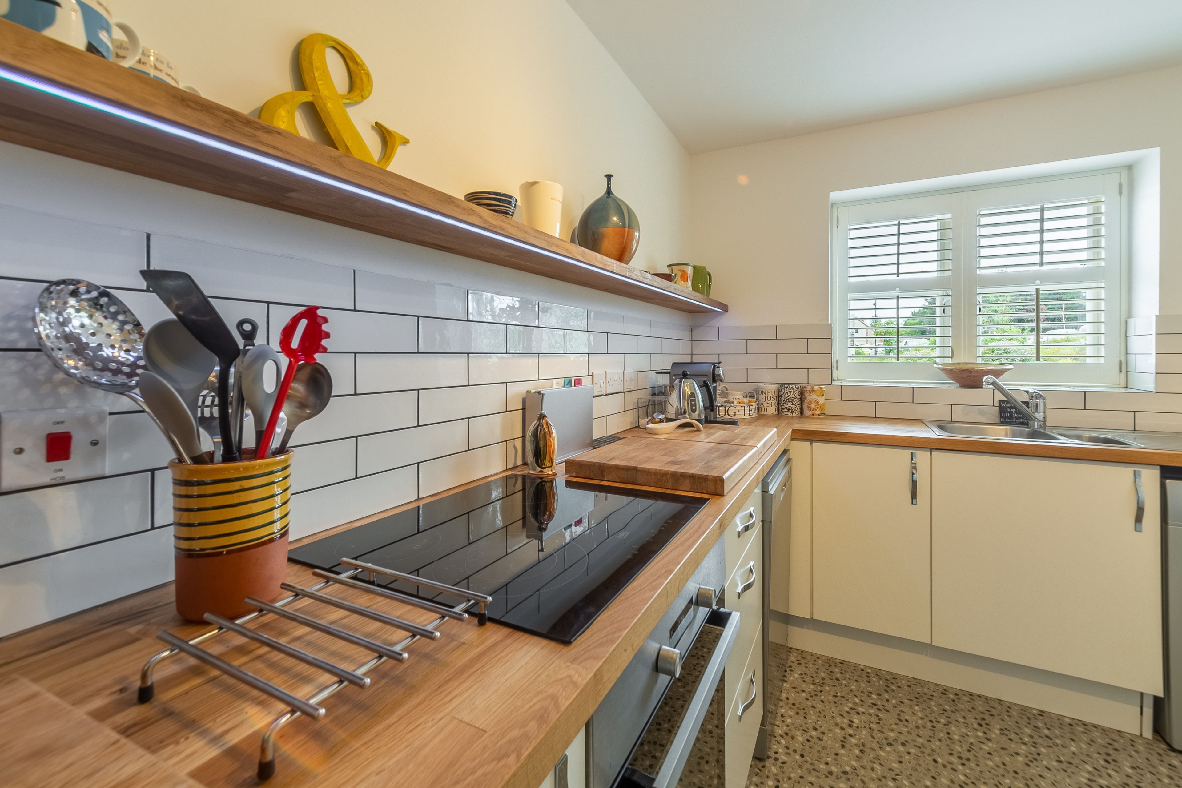 A modern kitchen with a black stovetop, wooden countertops, and white cabinets. There is a window with blinds, a sink, and a variety of kitchen utensils on the counter.