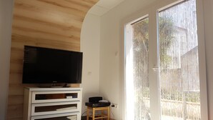 A modern living room with a flat-screen TV on a white cabinet, a small wooden table with electronic devices, and a sliding glass door leading to an outdoor area. - HOLIDAY RENTAL HOUSE WITH GARDEN IN PALAMOS (Palamos)