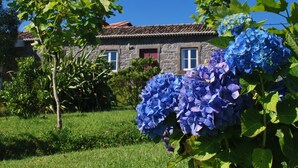 Un bâtiment en pierre, surmonté d’un toit de tuiles, entouré d’une végétation luxuriante et de fleurs d’hortensias bleues.