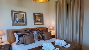 Un lit bien fait, avec des oreillers à rayures, une couverture marron et des serviettes posées sur le lit. Deux tableaux encadrés sont accrochés au mur au-dessus du lit, une lampe se trouve sur la table de chevet et un rideau est disposé à la fenêtre.