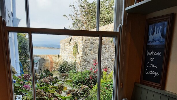 A wooden table with a bowl of fruit, a window with a view of a stone wall and greenery, and a 'Welcome to Curlew Cottage' sign. - 18th Century Cottage On Waterfront, Panoramic Sea Views, Courtyard Garden (Appledore, nr Bideford)