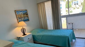 Ein Hotelzimmer mit zwei Betten, einem Nachttisch, einer Lampe, einem Gemälde und Blick auf Gebäude durch das Fenster.