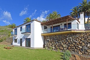 A modern two-story house with a stone wall foundation, large windows, and a balcony overlooking a grassy area. - Villa Vista Mar en Tijarafe (Tijarafe)