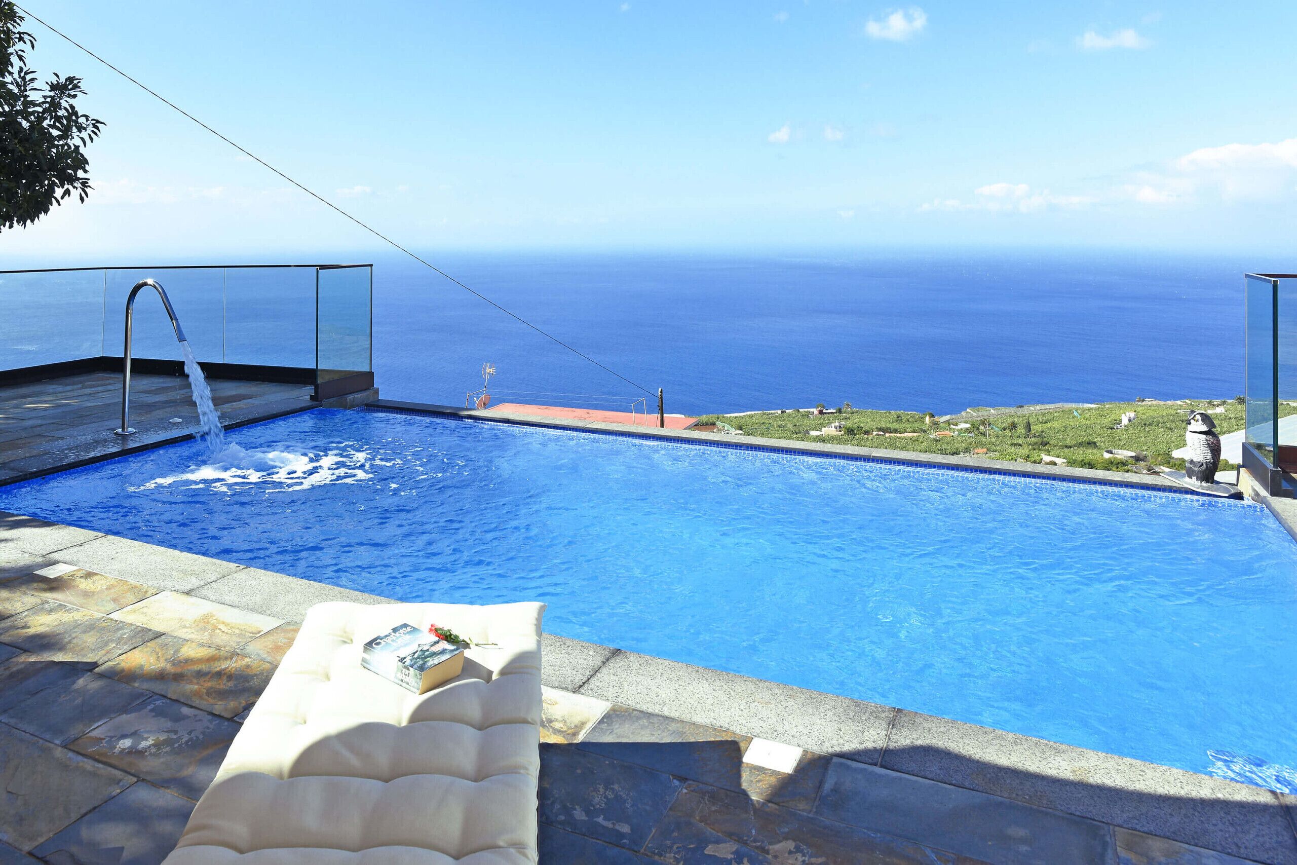 A swimming pool with a clear view of the ocean, a lounge area with a cushion, and a glass-enclosed deck.