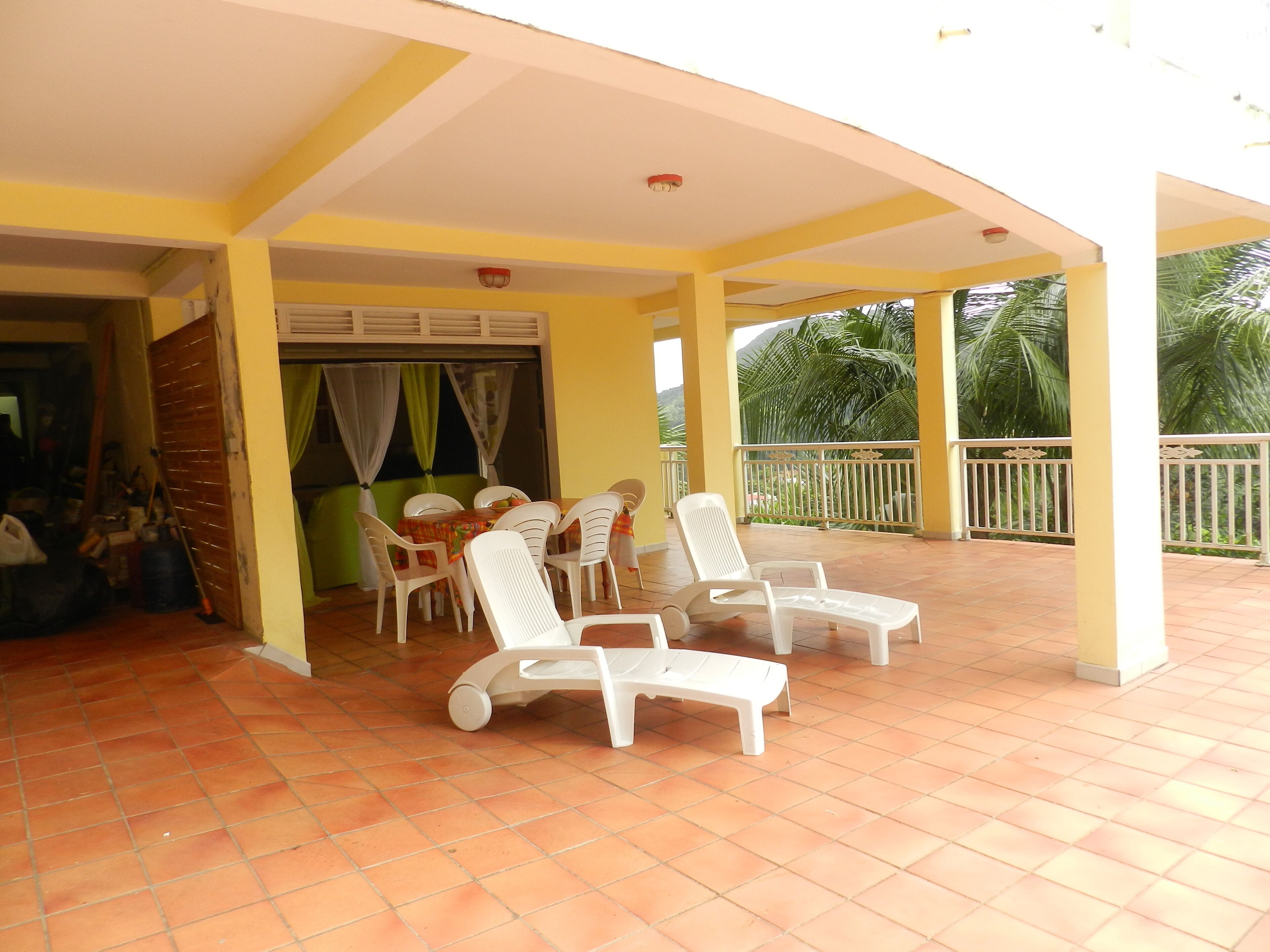 A covered outdoor seating area with white plastic chairs and a table, yellow walls, and a view of palm trees.