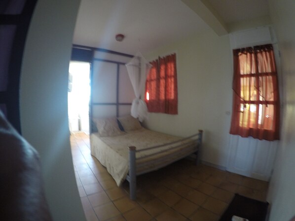 Una cama individual en una habitación con suelo de baldosas, una ventana con cortinas rojas y una puerta.