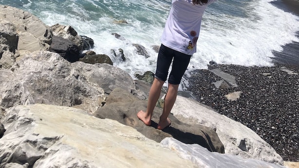 A person standing on a rocky shore with arms raised, overlooking a coastal area with waves crashing against the rocks and a pier extending into the water.