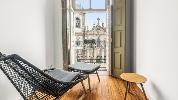 Un couloir étroit, avec une chaise en osier et un petit tabouret en bois, mène à un balcon doté d’une grille en fer forgé et offrant une vue sur un bâtiment historique.
