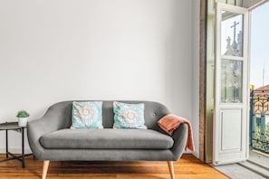 A grey sofa with patterned cushions, a small table with a plant, and a view of a building through an open door. - Almond Biscuit - Historic Center Harbor (Porto)