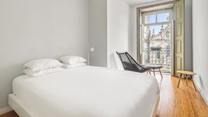 Une chambre à coucher avec un grand lit, une chaise, une petite table et une fenêtre donnant sur des immeubles.