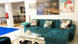 A teal sofa with floral and tropical patterned cushions, a glass coffee table with decorative items, and a floral-patterned rug.
