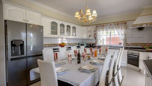 A modern kitchen with white cabinets, a stainless steel refrigerator, a dining table set for six, and a chandelier.