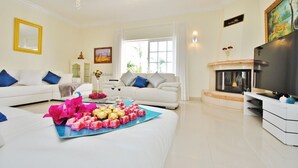 A living room with two white sofas, a coffee table with chocolates, a TV, and a fireplace.