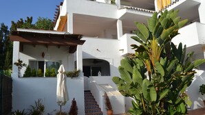 Un edificio blanco de dos pisos con una escalera de baldosas, plantas en macetas y una palmera de banano grande.