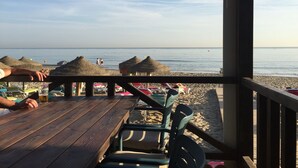 Terraza de madera con sillas y mesas con vista a una playa con sombrillas y cabañas de techo de paja.