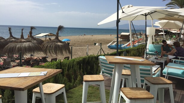 Un restaurante frente a la playa con mesas al aire libre, vista a la playa y sombrillas para dar sombra.