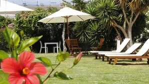 Un espace jardin aménagé avec une pelouse, des chaises longues, une table en bois et un parasol blanc.