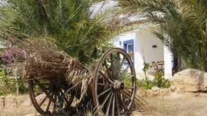 Un chariot en bois rustique, doté de grandes roues, est entouré d’herbes sèches et de rochers. En arrière-plan, on aperçoit un bâtiment blanc agrémenté de touches de bleu.