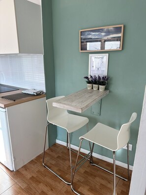 Una cocina pequeña con encimera de madera, dos taburetes blancos tipo bar, un letrero de bienvenida enmarcado y una pequeña planta en maceta sobre una estantería.