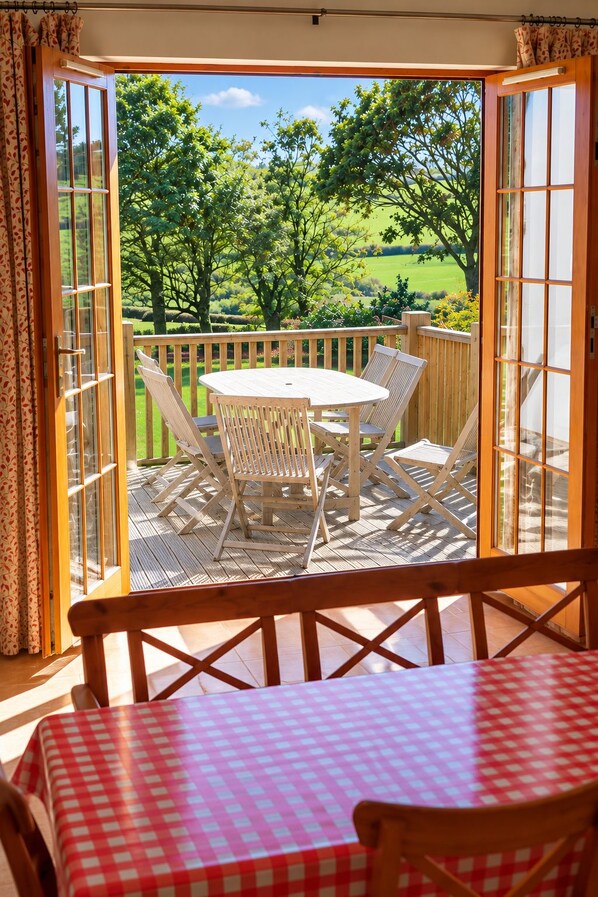 An open patio with a table and chairs, a view of a green landscape, and a wooden deck.