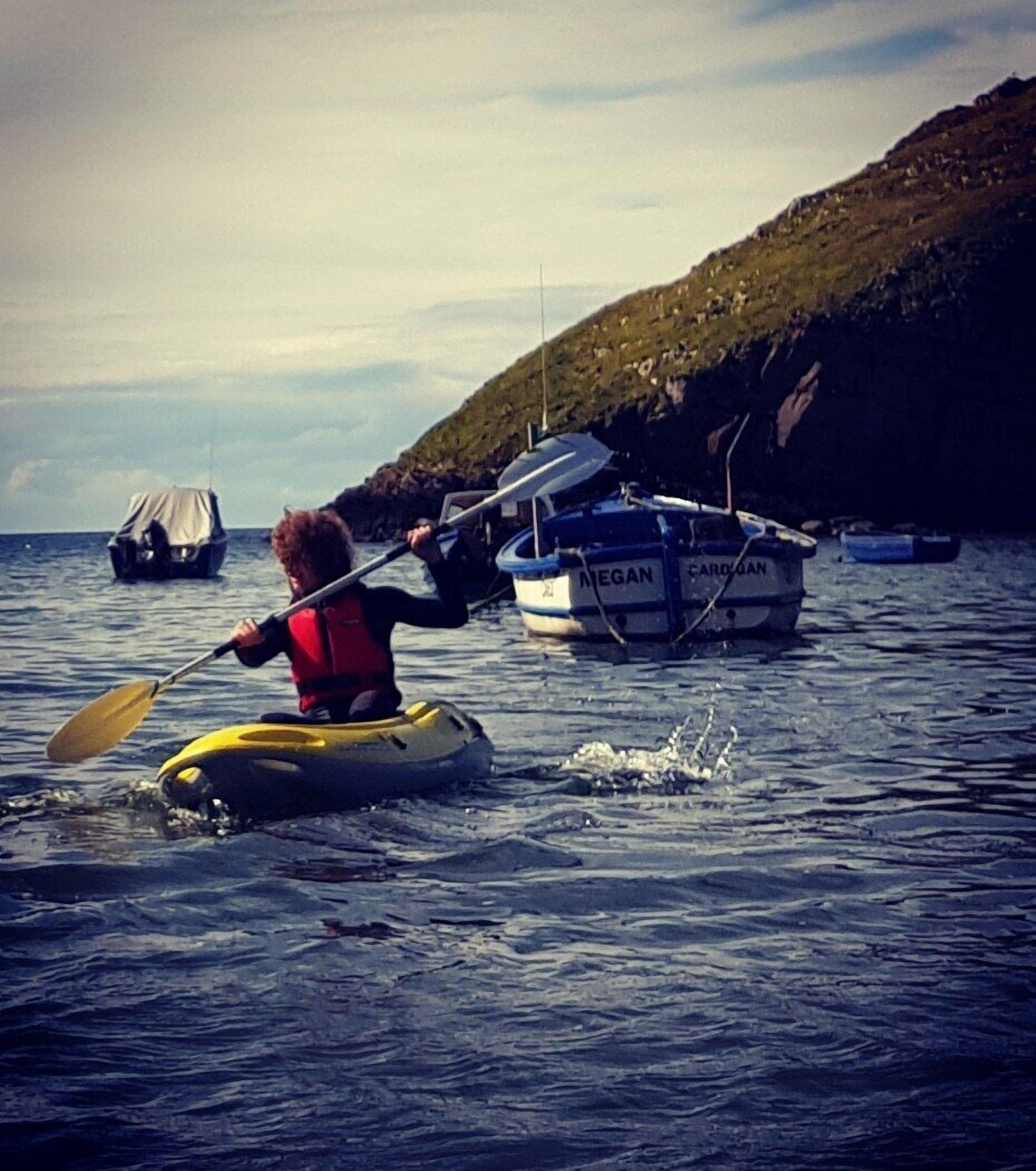 Une personne pratiquant le kayak sur une étendue d’eau, avec des bateaux en arrière-plan.