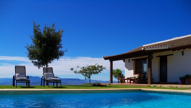 A pool with clear blue water, two lounge chairs, a tree, and a building with a tiled roof.