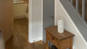 Un couloir avec une porte en bois, une petite table en bois sur laquelle est posé un objet blanc enroulé, et un escalier avec des rampes grises.