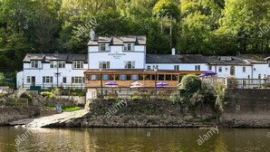 Un bâtiment situé au bord de la rivière, avec une façade blanche, une terrasse en bois dotée de parasols et un mur en pierre.