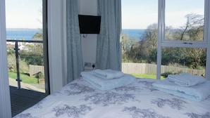A bedroom with a bed, a TV mounted on the wall, and a view of the sea. - PRISTINE APARTMENT, 4 MIN WALK TO PRIVATE BEACH, STUNNING SEA VIEWS. (Duporth)