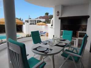 Eine Terrasse mit einem runden Esstisch, umgeben von türkisfarbenen Stühlen, einem Grill und einem Pool im Hintergrund.