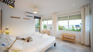 A hotel room with a bed, bedside lamps, a wooden bench, a window with curtains, and a view of greenery outside. - Beachside Luxury Villa Private Heated Pool & Sea Views in Las Chapas (Marbella)