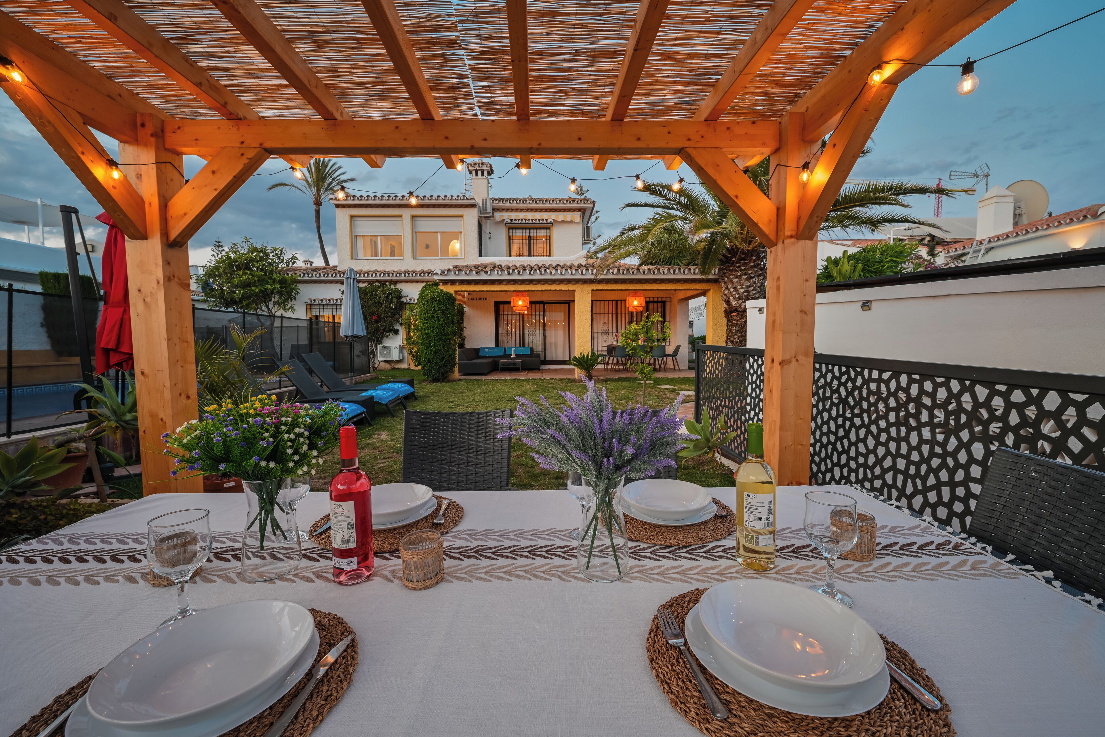 Een buitenplaats met een pergola, tafels gedekt met witte tafellakens, en een uitzicht op een huis op de achtergrond.