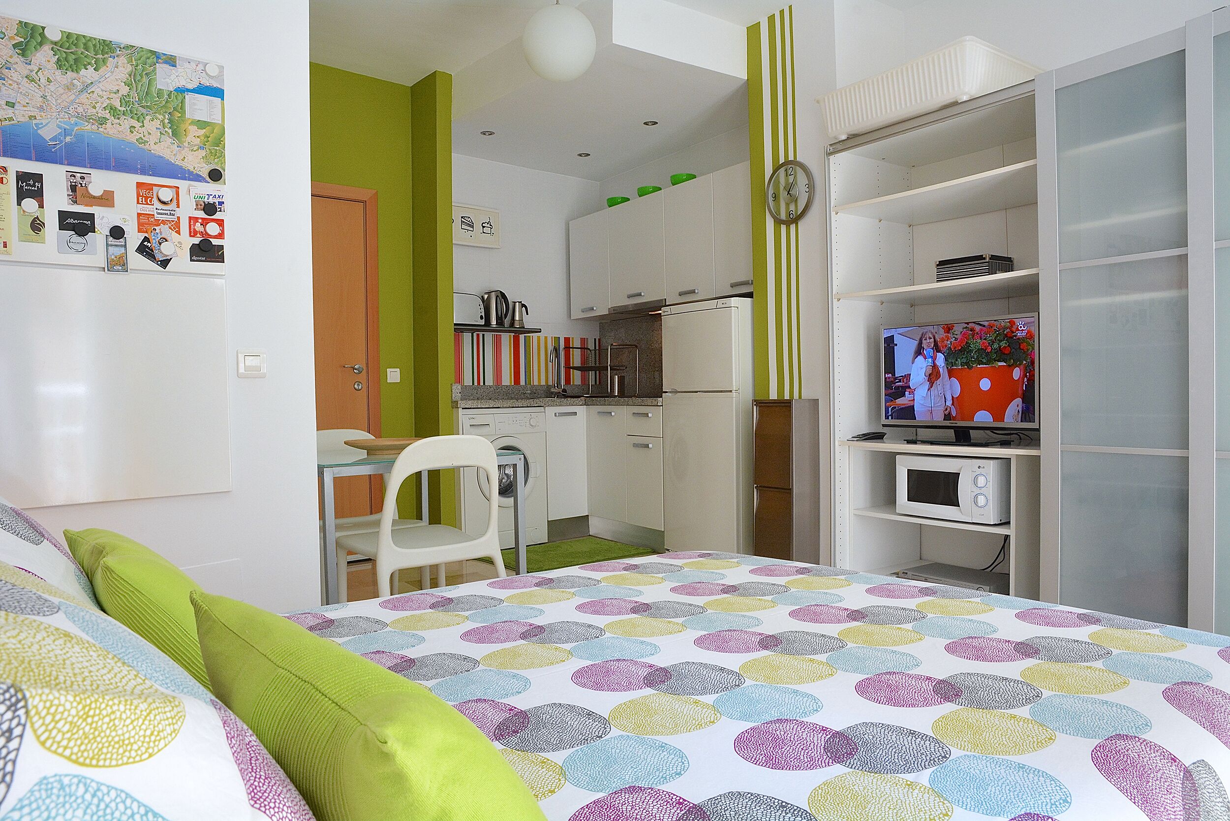 A modern bedroom with a bed, green pillows, a colorful bedspread, a kitchenette, a TV, and a map on the wall.