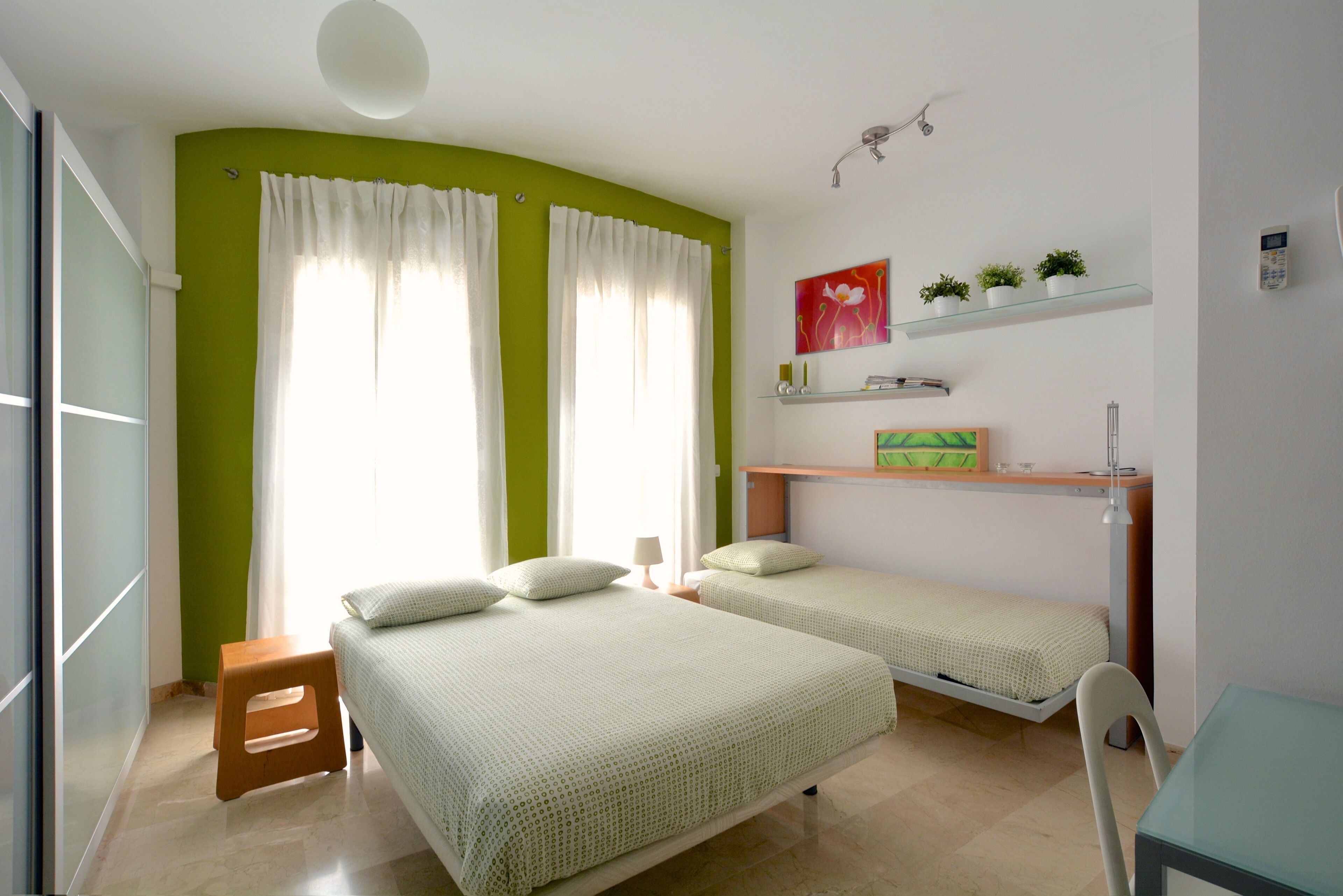 A room with two beds, a small table, and a chair. There are shelves with plants and a painting on the wall.