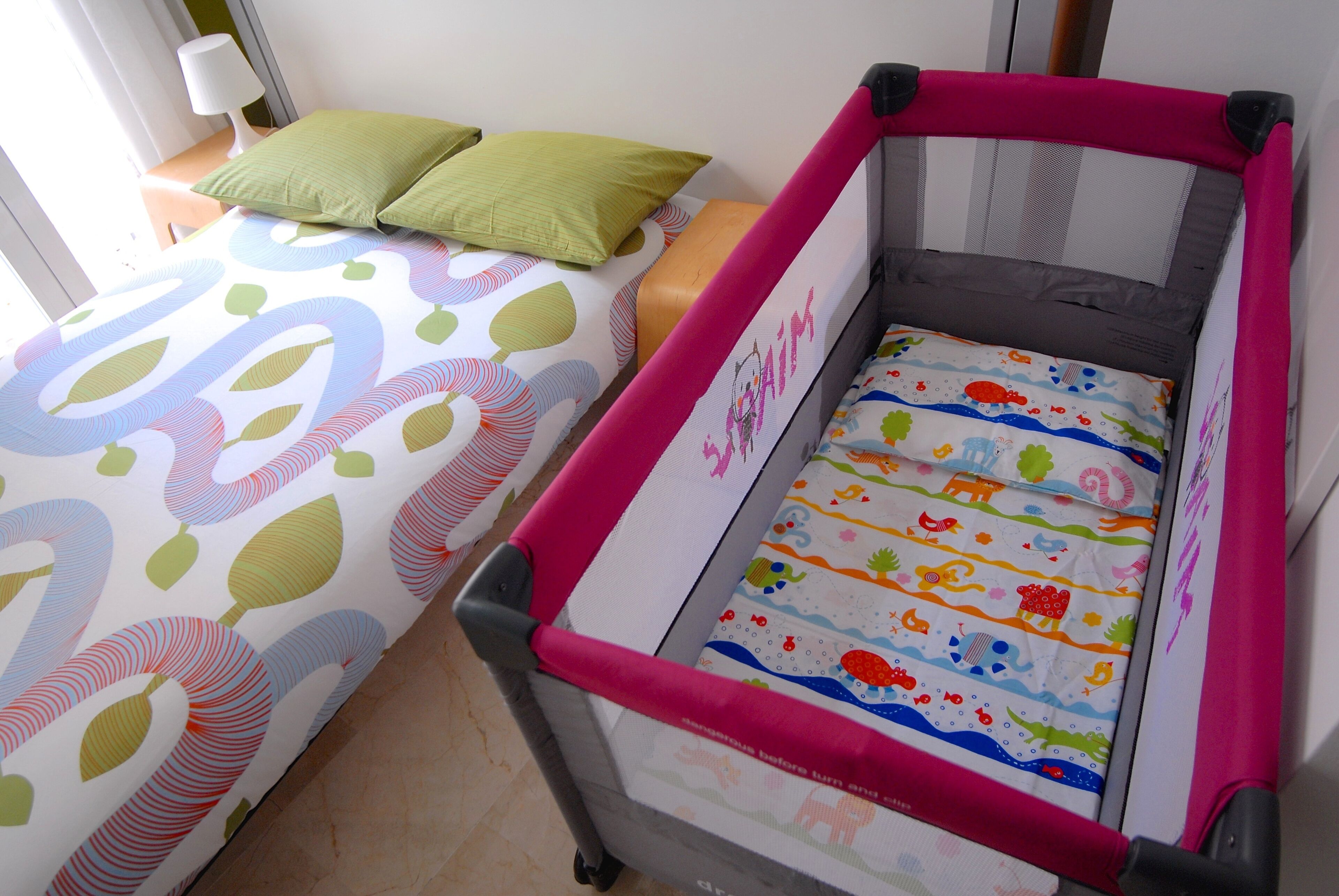 Una corralita infantil con un diseño colorido, una cama con ropa de cama estampada y un almohadón verde sobre la cama.