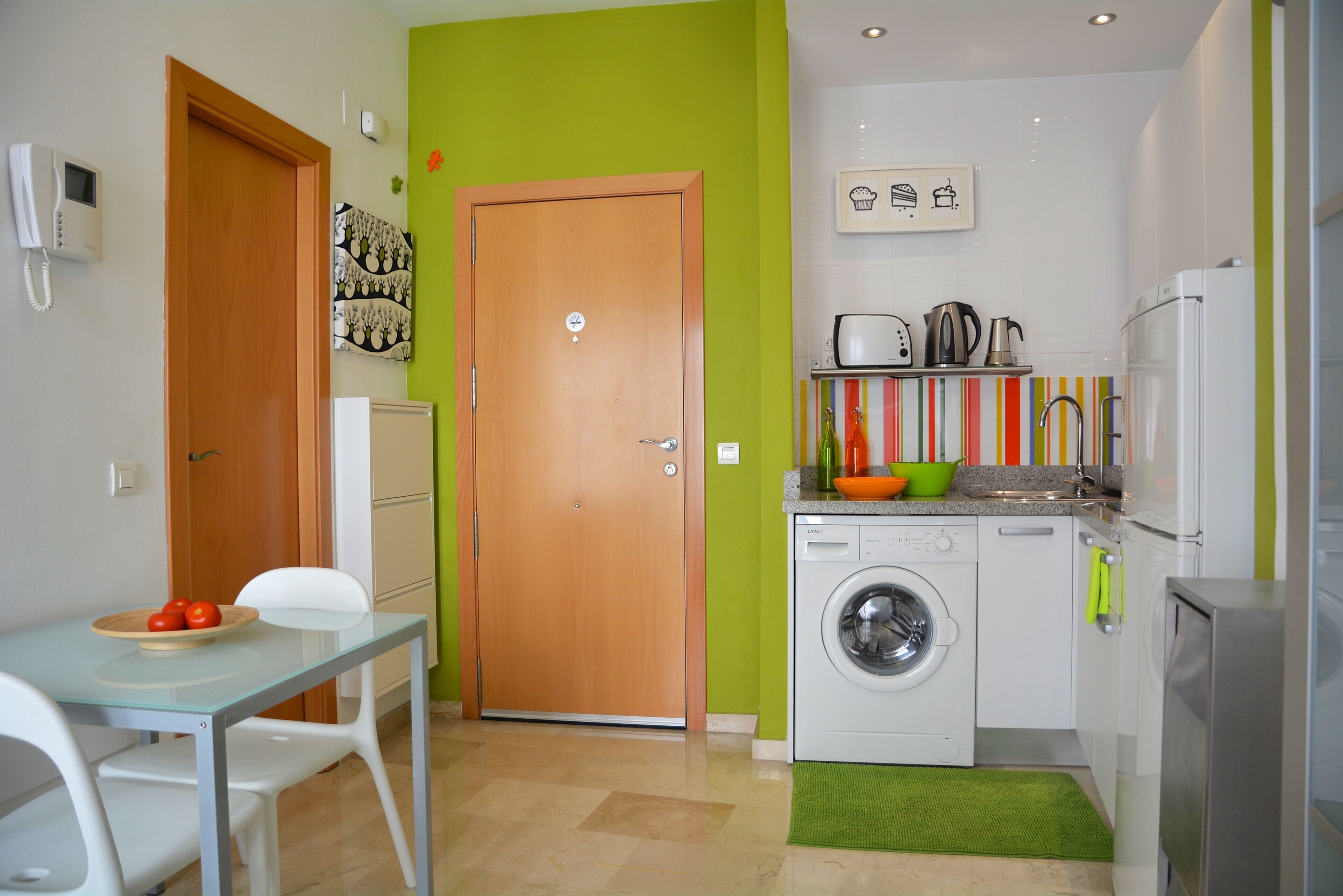 A compact kitchen with a washing machine, a small dining table with chairs, and a door with a light switch.