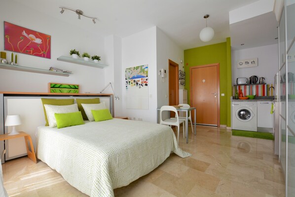 A bedroom with a bed, bedside table, and a small desk with a chair. There is a kitchen area with a washing machine and a refrigerator.