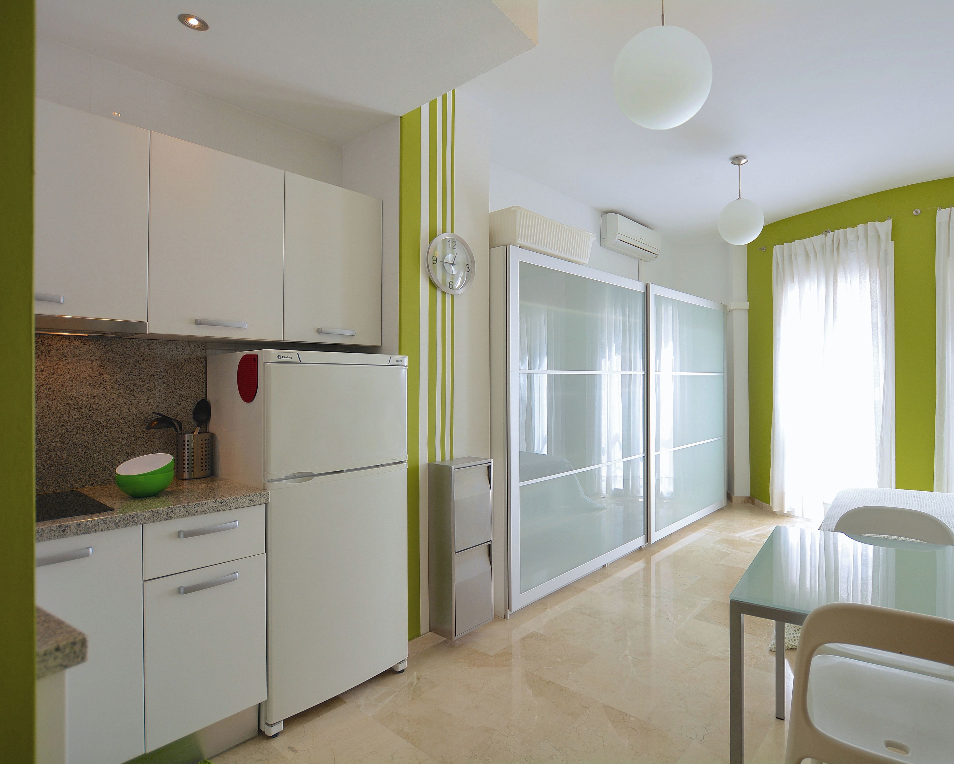 Cozinha moderna com armários brancos, parede com listras verdes, geladeira e área de jantar com mesa e cadeiras de vidro.