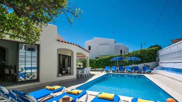 A pool area with blue lounge chairs and umbrellas, a clear blue sky, and white buildings with red-tiled roofs. - Algarve, Casa Julio, Albufeira Town, Beside The Strip (Albufeira)